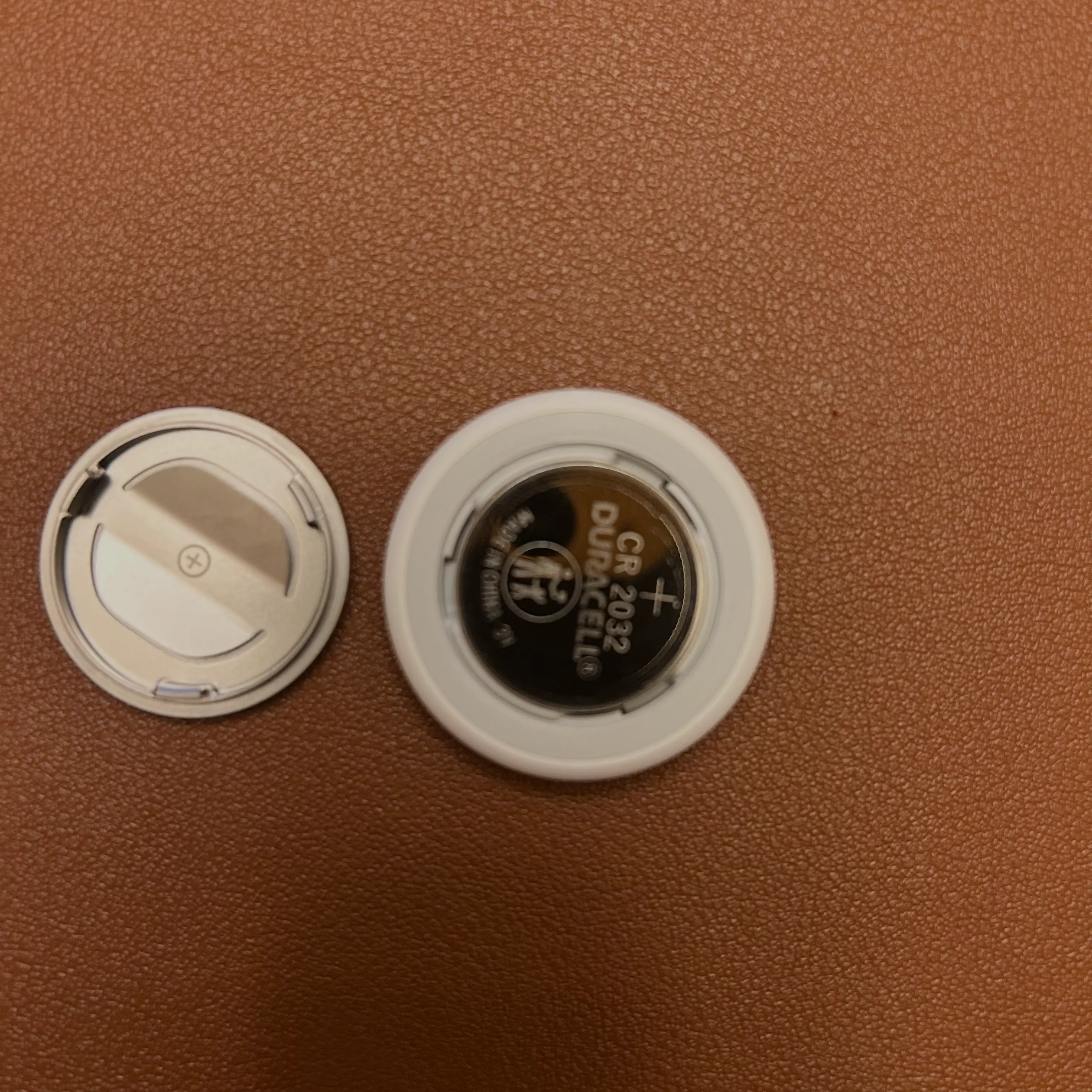 The AirTag with a new CR2032 battery inserted into the battery compartment, next to the metal cover that has been removed. TThe AirTag with a new CR2032 battery inserted into the battery compartment, next to the metal cover that has been removed.