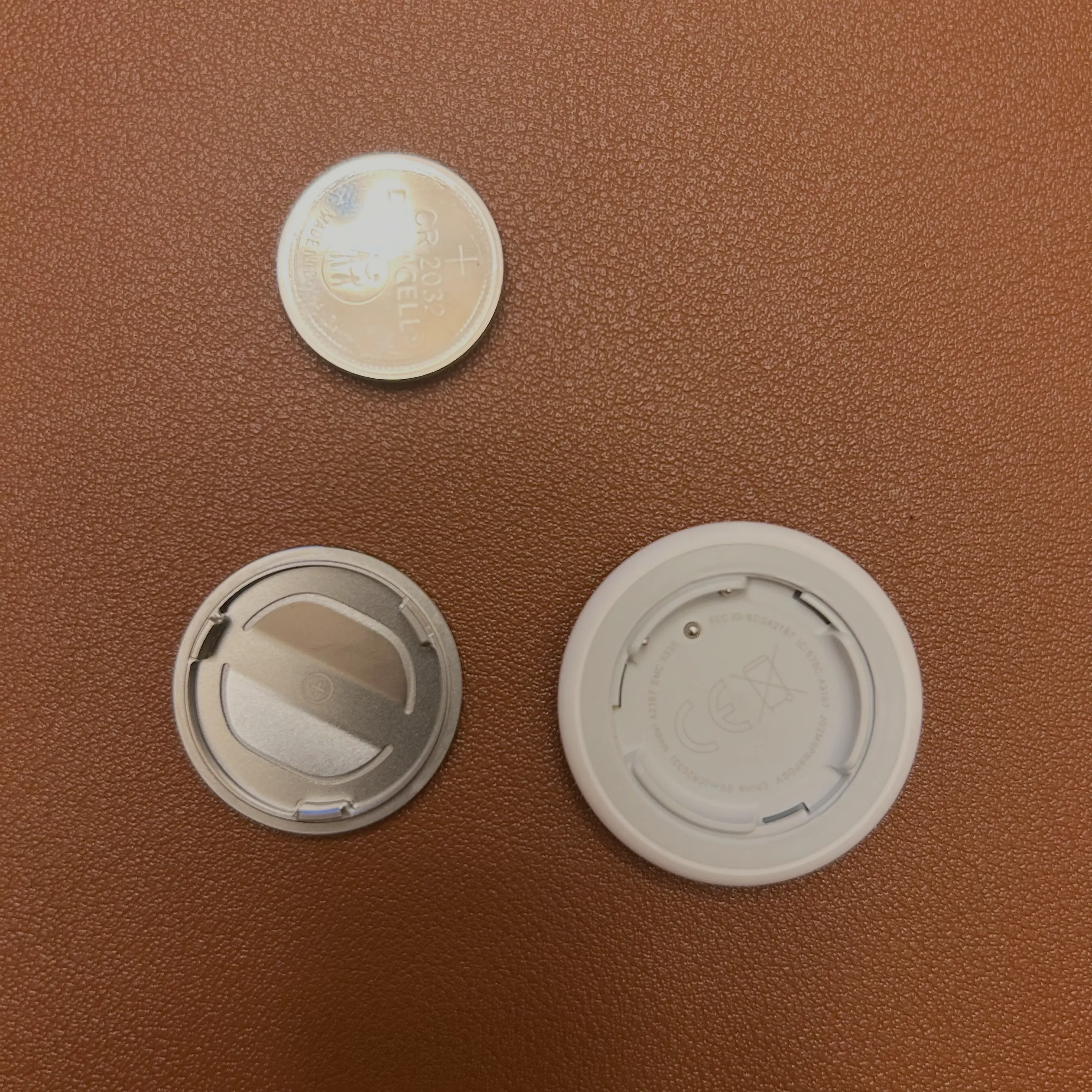 The AirTag's components laid out on a brown surface, including the metal back cover, the removed CR2032 battery, and the AirTag's plastic casing. The AirTag's components laid out on a brown surface, including the metal back cover, the removed CR2032 battery, and the AirTag's plastic casing.